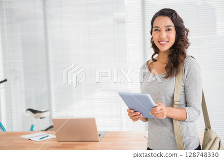 Asian woman smiling while holding tablet in bright office at desk with laptop and bicycle wheel 128748330