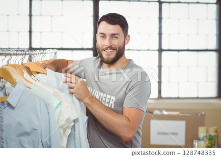 Male volunteer sorting donated shirts on wooden hangers beside donation boxes at warehouse 128748335