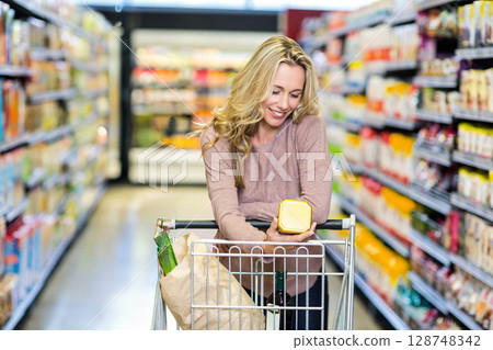 Mid adult woman pushing shopping cart with grocery bag and wine reading jar label at supermarket 128748342