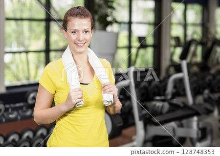 Female gym-goer standing in gym wearing yellow sportswear and holding white towel by dumbbell rack Female gym-goer standing in gym wearing yellow sportswear and holding white towel by dumbbell rack 128748353