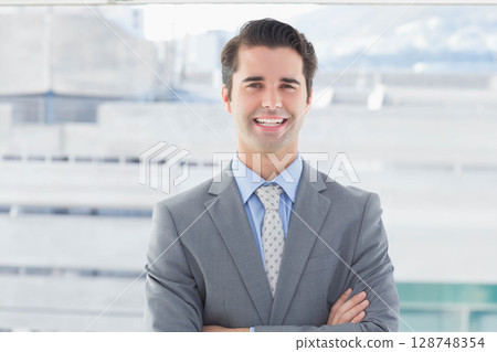 Businessman standing smiling arms crossed near concrete railing on rooftop in grey suit and tie Businessman standing smiling arms crossed near concrete railing on rooftop in grey suit and tie 128748354