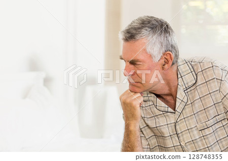 Senior man sitting on bed near window blinds wearing plaid pajama top in bedroom, copy space Senior man sitting on bed near window blinds wearing plaid pajama top in bedroom, copy space 128748355
