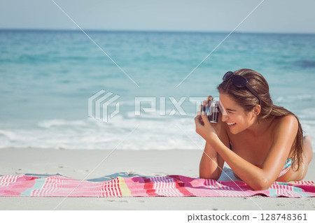 Woman in her twenties wearing floral bikini lying on beach towel, holding retro camera, copy space Woman in her twenties wearing floral bikini lying on beach towel, holding retro camera, copy space 128748361