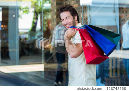 Mid-adult man standing at retail storefront, carrying three paper bags, smiling back, copy space 128748368