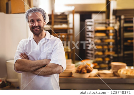 Male baker standing with arms crossed in bakery beside steel counter with baskets, copy space 128748371