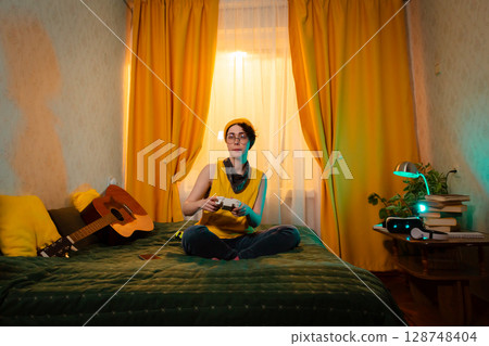 Calm teenage girl enjoying gaming after school in her room. Holding gamepad, leaning on pillows. Concept of comfort, tech and youth identity. Wide shot Calm teenage girl enjoying gaming after school in her room. Holding gamepad, leaning on pillows. Concept of comfort, tech and youth identity. Wide shot 128748404