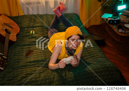 Caucasian teen girl with game controller smiling and relaxing. Cozy bed and warm room design. Concept of joyful rest after school. Top view 128748406