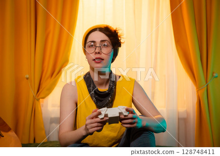 Portrait of concentrated teen girl dressed in loose clothes playing with game controller. Cozy bedroom with yellow curtains. Concept of relaxation and screen time. Portrait of concentrated teen girl dressed in loose clothes playing with game controller. Cozy bedroom with yellow curtains. Concept of relaxation and screen time. 128748411