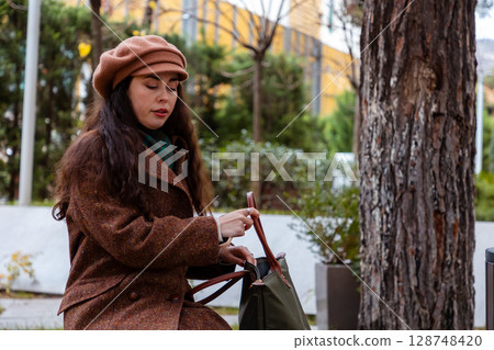 Stylish Caucasian woman resting on a park bench with her purse during a calm city walk. Relaxation, street fashion, and everyday female lifestyle in urban surroundings. Stylish Caucasian woman resting on a park bench with her purse during a calm city walk. Relaxation, street fashion, and everyday female lifestyle in urban surroundings. 128748420