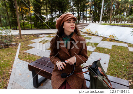 Young Caucasian woman in a coat sits alone on a bench in the city, relaxing with her handbag beside her. Urban lifestyle, weekend break, and peaceful outdoor moment. 128748421