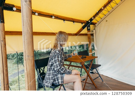 woman sitting inside glamping tent holding cigarette and phone on wooden chair at rustic table with casual outfit under yellow canopy, outdoor travel, countryside lifestyle, digital detox 128750309
