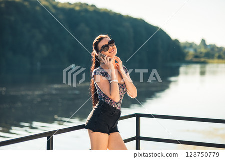 The girl with a long braided ponytail is cheerfully talking on the phone by the lake 128750779