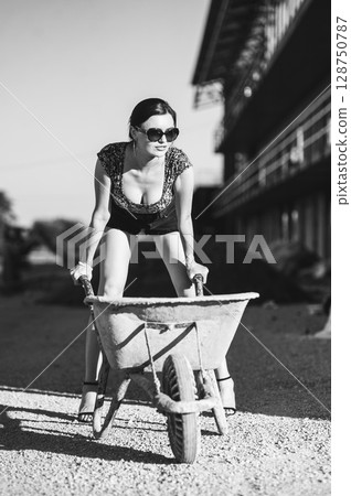 A young girl confidently poses by an old wheelbarrow in front of an unfinished building 128750787