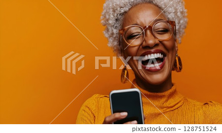 Joyful mature woman with gray curly hair laughing while using smartphone. Natural lifestyle portrait against vibrant orange background showcasing authentic emotion 128751524
