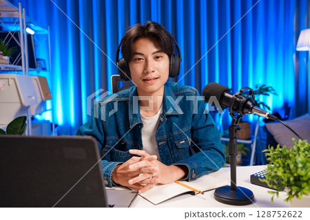 Man podcaster influencer blogger smiling while broadcasting his live audio podcast in studio using headphones. Male radio host making podcast or interview. 128752622