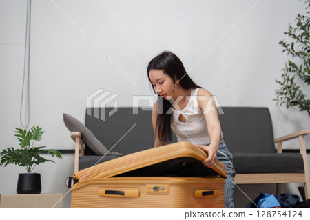 Young Woman Packing Suitcase for Travel Adventure in Modern Living Room Setting with Stylish Decor and Casual Clothing Young Woman Packing Suitcase for Travel Adventure in Modern Living Room Setting with Stylish Decor and Casual Clothing 128754124