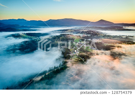 Stunning aerial view of sunrise over mountainous landscape, with thick fog blanketing valleys and hills. Sun's golden rays pierce through mist, casting serene glow over the entire scene. 128754379
