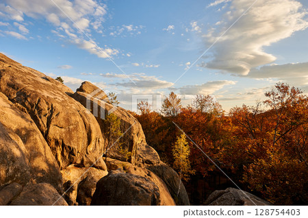 Golden hues of setting sun illuminate rocky formations and autumn foliage, casting warm glow on scene. Vibrant oranges and reds of trees below. Dovbush Rocks, Carpathian mountains, Ukraine, Europe. 128754403