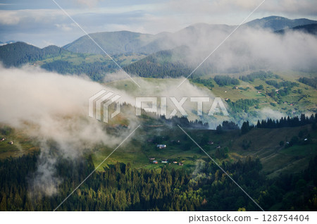 Picturesque scene of mountain valley blanketed in mist, with patches of green pastures and scattered houses. Clouds drift gently over forested hills, creating serene and tranquil atmosphere. 128754404