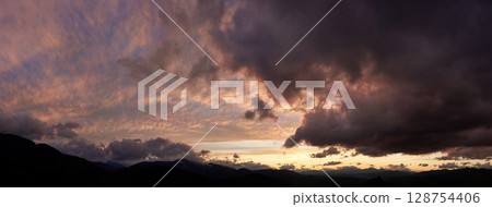 Dramatic sunset scene with dark, brooding clouds casting shadows over sky. Horizon glows with warm hues of orange and pink, contrasting against silhouettes of mountains, creating moody atmosphere. 128754406