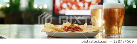 Super Bowl game party, glass of beer and plate of chips with salsa next to television screen 128754509
