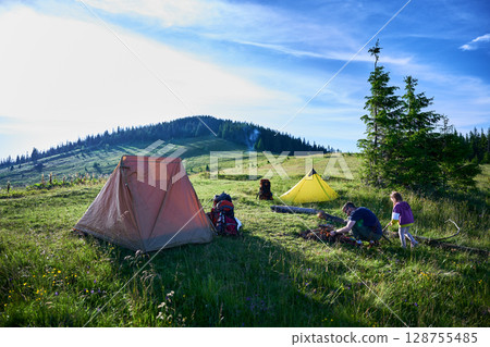 Picturesque mountain campsite on grassy hill with tents. Camper and child tend to campfire, surrounded by backpacks and gear. Rolling hills, evergreens, clear blue sky. Peaceful outdoor adventure. 128755485