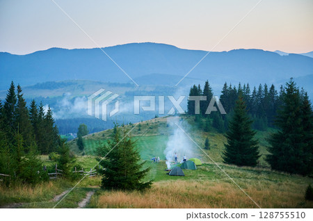 Aerial view campsite in mountains, with several colorful tents on grassy clearing. Campers relax under clear blue sky, surrounded by tall evergreens and panoramic views of distant peaks. 128755510
