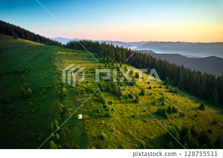 Aerial view of campsite on grassy hillside, with tents scattered amidst lush greenery and small trees. Golden hues of sunrise. In distance, layers of rolling mountains fade into horizon. Aerial view of campsite on grassy hillside, with tents scattered amidst lush greenery and small trees. Golden hues of sunrise. In distance, layers of rolling mountains fade into horizon. 128755511