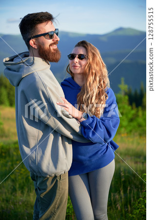 Portrait of couple stand close together in scenic meadow with mountains in background. Man and woman wearing sunglasses. Serene and affectionate moment amidst natural beauty. 128755515