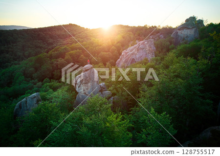 Aerial view of climber standing atop rock formation at Dovbush Rocks in Carpathian mountains, Ukraine. Sun sets, casting warm glow over lush, green forest, distant hills, creating breathtaking view. 128755517