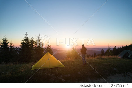 Tranquil sunrise over mountain campsite, with vibrant yellow tent and lone camper standing nearby. Tall evergreens silhouetted against soft morning light, with rolling hills fading into horizon. 128755525