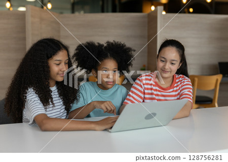 Diverse group of kids three girls happy looking at computer screen together, modern workspace. They are seated at white table while sharing ideas and looking at laptop in classroom school. 128756281