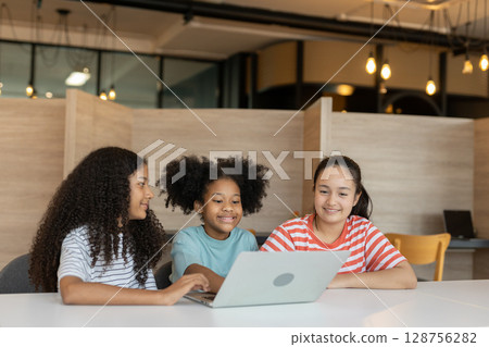 Diverse group of kids three girls happy looking at computer screen together, modern workspace. They are seated at white table while sharing ideas and looking at laptop in classroom school. Diverse group of kids three girls happy looking at computer screen together, modern workspace. They are seated at white table while sharing ideas and looking at laptop in classroom school. 128756282