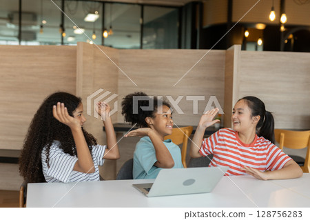 Diverse group of kids three girls happy looking at computer screen together, modern workspace. They are seated at white table while sharing ideas and looking at laptop in classroom school. Diverse group of kids three girls happy looking at computer screen together, modern workspace. They are seated at white table while sharing ideas and looking at laptop in classroom school. 128756283