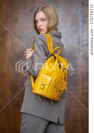 A stylish young woman, long blonde hair, poses in a grey suit, holding a vibrant yellow leather backpack against a warm wooden background A stylish young woman, long blonde hair, poses in a grey suit, holding a vibrant yellow leather backpack against a warm wooden background 128756314