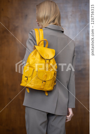 A stylish young woman, long blonde hair, poses in a grey suit, holding a vibrant yellow leather backpack against a warm wooden background A stylish young woman, long blonde hair, poses in a grey suit, holding a vibrant yellow leather backpack against a warm wooden background 128756315