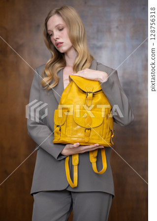 A stylish young woman, long blonde hair, poses in a grey suit, holding a vibrant yellow leather backpack against a warm wooden background A stylish young woman, long blonde hair, poses in a grey suit, holding a vibrant yellow leather backpack against a warm wooden background 128756318