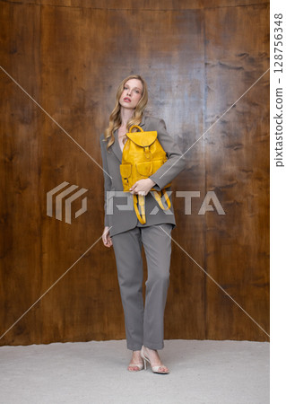 A stylish young woman, long blonde hair, poses in a grey suit, holding a vibrant yellow leather backpack against a warm wooden background 128756348
