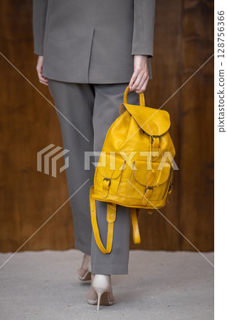 A stylish young woman, poses in a grey suit, holding a vibrant yellow leather backpack against a warm wooden background 128756366