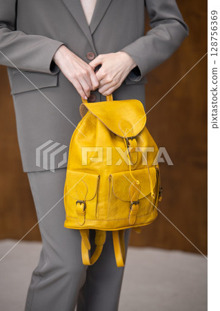 A stylish young woman, poses in a grey suit, holding a vibrant yellow leather backpack against a warm wooden background 128756369