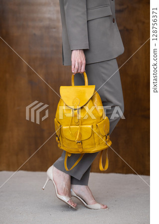 A stylish young woman, poses in a grey suit, holding a vibrant yellow leather backpack against a warm wooden background 128756371