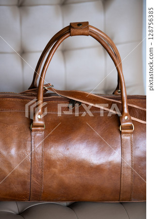 A vintage-style brown leather duffel bag rests on a cream-colored tufted chair against a blurred wooden and grey background. Part photo 128756385