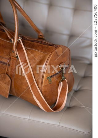 A vintage-style brown leather duffel bag rests on a cream-colored tufted chair against a blurred wooden and grey background. Part photo A vintage-style brown leather duffel bag rests on a cream-colored tufted chair against a blurred wooden and grey background. Part photo 128756400