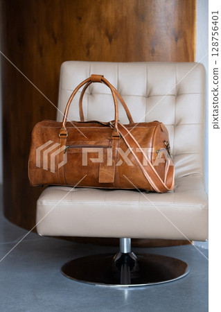 A vintage-style brown leather duffel bag rests on a cream-colored tufted chair against a blurred wooden and grey background A vintage-style brown leather duffel bag rests on a cream-colored tufted chair against a blurred wooden and grey background 128756401