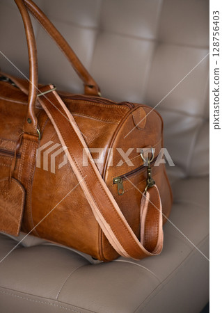 A vintage-style brown leather duffel bag rests on a cream-colored tufted chair against a blurred wooden and grey background. Part photo A vintage-style brown leather duffel bag rests on a cream-colored tufted chair against a blurred wooden and grey background. Part photo 128756403