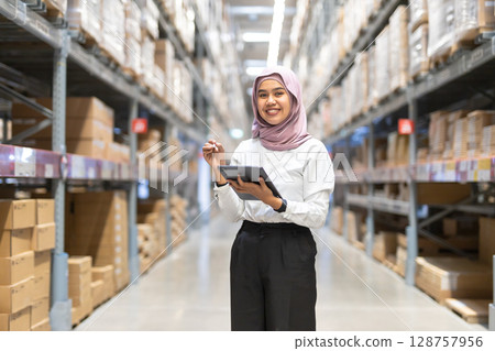 Young muslim women wearing hijab smiles while holding tablet checking goods stock inspacious warehouse. Female islamic worker at large storage facility. 128757956