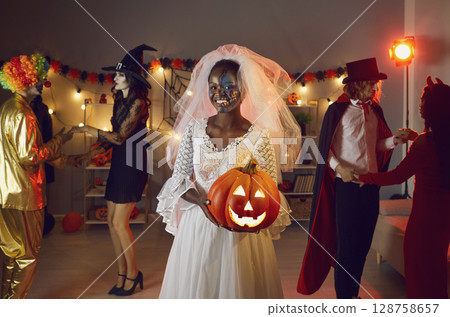 Happy black woman dressed as Dead Bride with her jack-o-lantern at Halloween party Happy black woman dressed as Dead Bride with her jack-o-lantern at Halloween party 128758657