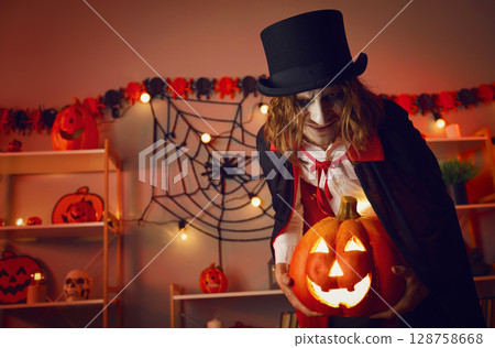 Spooky portrait of vampire man standing in room in dark with glowing jack-o-lantern in his hands. 128758668