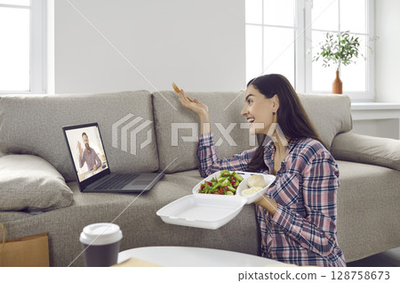 Happy woman talking to friend on video call while eating takeaway meal on lunch break 128758673
