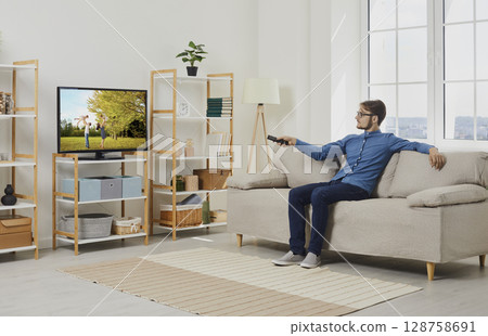 Young man relaxing on sofa in living room and watching film on large screen of modern TV 128758691